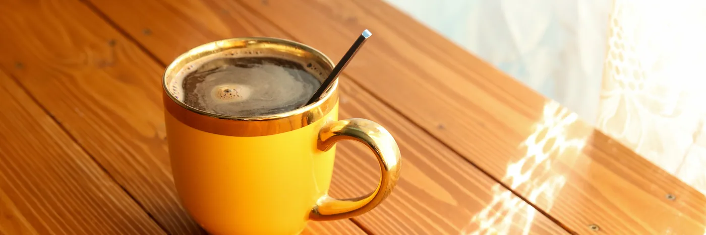 A yellow ceramic cup of black coffee with a spoon, standing on a wooden table in low morning sunlight.