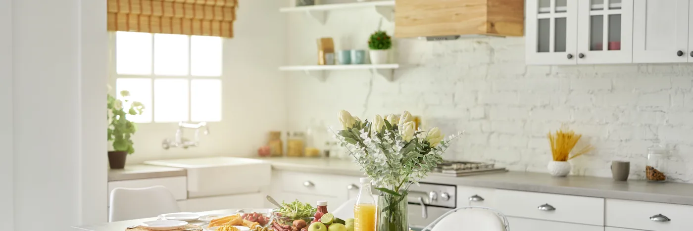 A bright white kitchen with a window over the sink, open shelving and a vase of flowers beside a laid table.