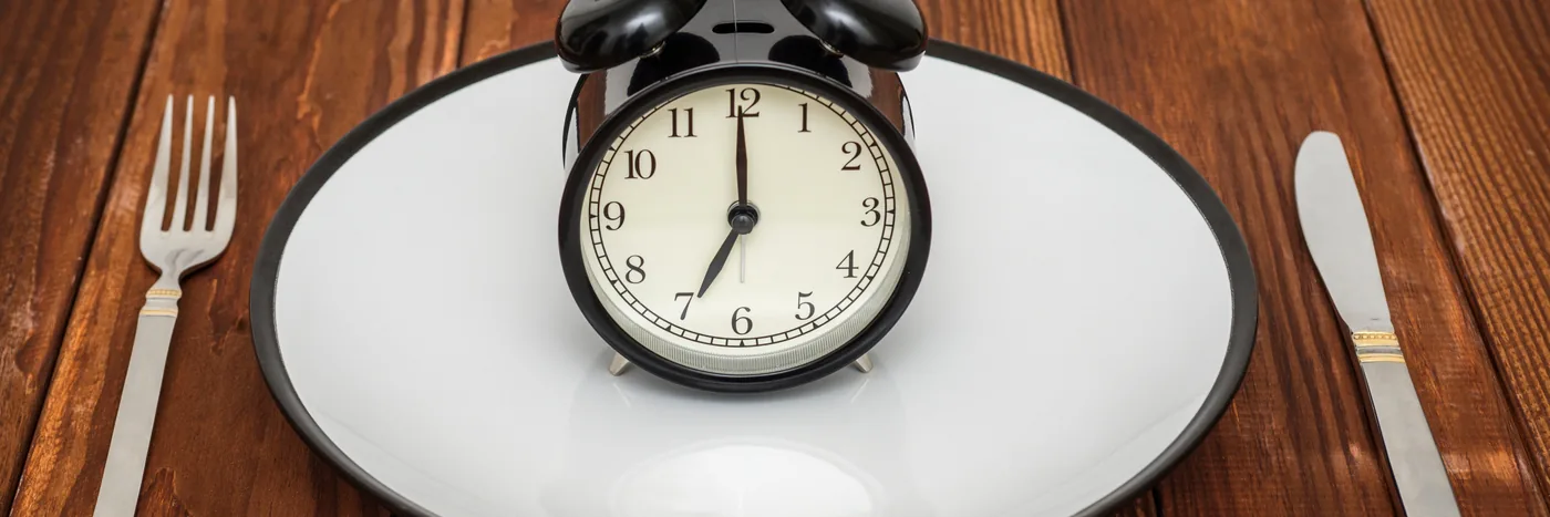A black alarm clock sitting on a white dinner plate between a fork and a knife on a dark wooden table.