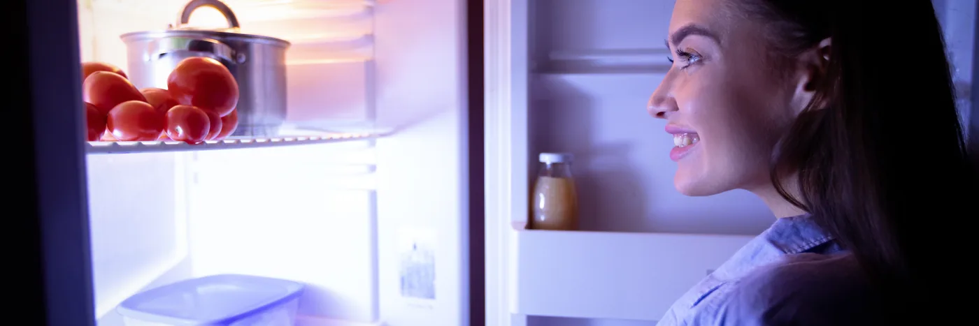 A woman in profile lit only by the open door of a refrigerator in a dark kitchen at night.
