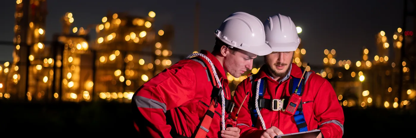 Two workers in hard hats and red overalls looking at a tablet at night, an industrial plant lit up behind them.