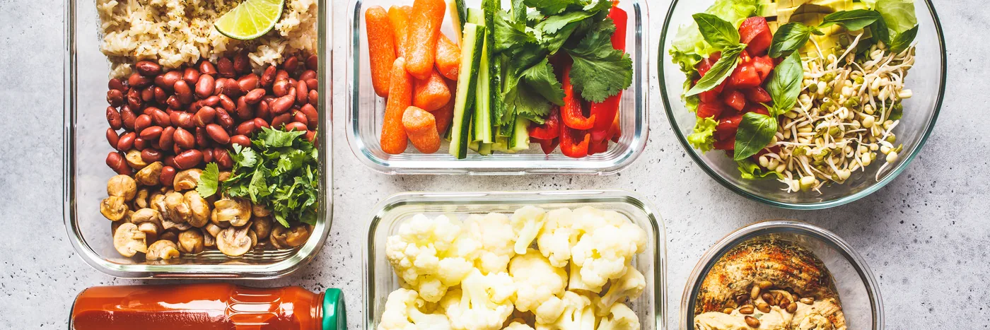 Glass meal-prep containers of beans, cut vegetables, salad and rice photographed from directly above.